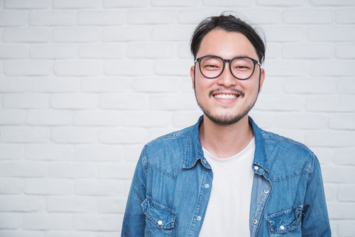 man standing in front of white brick wall while smiling and wearing glasses and denim jacket
